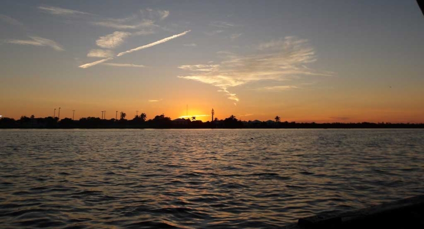 The sky appears in gentle colors as the sun sets behind a body of water and a shoreline in the distance. 